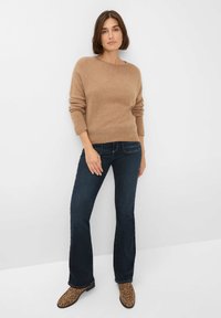 Beige gebreide trui met lange mouwen, gecombineerd met donkerblauwe wijde jeans en bruine enkellaarsjes met panterprint. Minimalistische achtergrond.