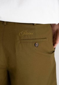 Brune shorts med en glatt tekstur, med en knappelukking og gul brodert "Forbudt" på baklommen.