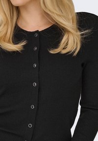 Femme blonde portant un cardigan noir côtelé avec des boutons noirs bordés d'argent, vue du cou à la taille sur un fond blanc.
