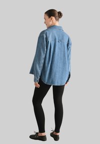 Kobieta stoi, ubrana w luźną niebieską dżinsową kurtkę, czarne legginsy, czarne sznurowane buty, z włosami w kok, zwrócona bokiem.