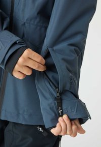 Giacca impermeabile blu navy con polsino con zip, che mostra una texture liscia e un dettaglio del logo in rilievo. Presenta un design del polso regolabile per una vestibilità su misura.