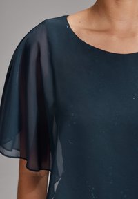 Femme portant un haut en tissu transparent bleu foncé avec des manches courtes et larges ainsi qu’un large col rond.