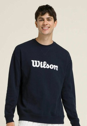 Giovane sorridente che indossa una felpa nera Wilson e pantaloni bianchi davanti a uno sfondo chiaro e uniforme.