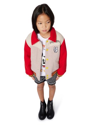 Giacca varsity rossa e beige con bottoni, dettagli gialli e ricami. Indossata sopra una camicia bianca e pantaloni shorts in denim con stivali neri.