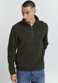 Pullover in pile verde scuro con collo alto, chiusura a zip quarti e discreto dettaglio del logo sul petto. Texture morbida e vestibilità comoda.