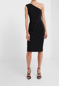 Svart, enaxlad klänning med figurnära design, knälång fåll och subtil silverdetalj vid midjan. Slitstarkt tyg med lätt lyster.