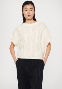 Chunky Strickpullover in sanftem Creme mit kurzen Ärmeln und lockerem Schnitt. Verfügt über ein strukturiertes Zopfmuster und einen gerippten Saum. Kombiniert mit dunkel gestreiften Hosen.