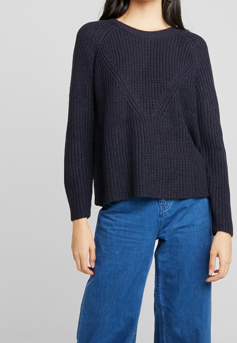 Ciemnogranatowy sweter z dzianiny o luźnym kroju, z szerokim dekoltem i prążkowaną teksturą. W zestawie z niebieskimi spodniami o szerokich nogawkach.