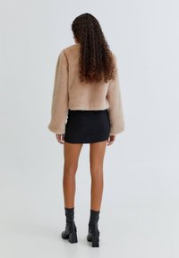 Ljusbrun pälsjacka med långa ärmar, bärs över en svart minisär. Modellen har lockigt hår och svarta klackstövlar, visad bakifrån.