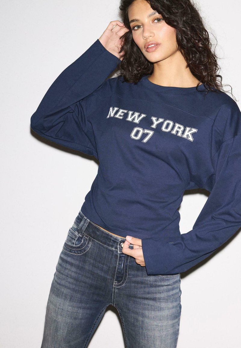 Donna con capelli ricci che indossa una maglietta a maniche lunghe blu navy con la scritta "NEW YORK 07" e jeans blu sbiaditi, posa con una mano alzata.