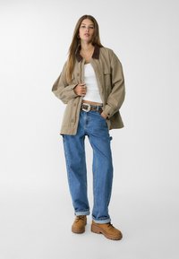 Beige oversized jakke med brun krave, hvid tanktop, blå løse jeans og brune snørebåndsstøvler. Accentbælte med nitter.