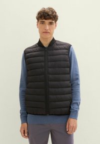 Schwarze Daunenweste mit genähten Abschnitten, vorne mit Reißverschluss und geripptem Kragen, getragen über einem blauen Langarmshirt. Modell steht vor einem hellen Hintergrund.