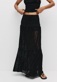 Jupe maxi en dentelle noire avec un design en couches, dotée d'une taille ajustée et d'une texture transparente. Portée avec un haut noir et des sandales.