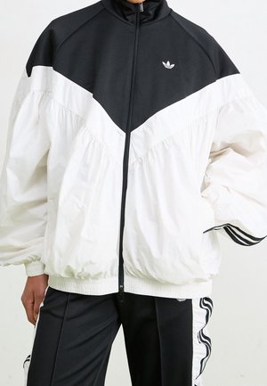 Persona che indossa una giacca da allenamento Adidas nera e bianca con zip frontale e pantaloni da allenamento neri abbinati con strisce bianche.