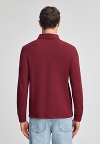 Bordeauxfarbene Langarm-Polohemd mit Kragen, strukturiertem Stoff und gerippten Bündchen, kombiniert mit hellblauen Jeans.