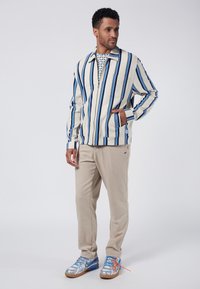 Randig zip-jacka i krämfärg med blå linjer, beige byxor och sneakers med blå accenter. Avslappnad look med distinkt vertikala ränder.