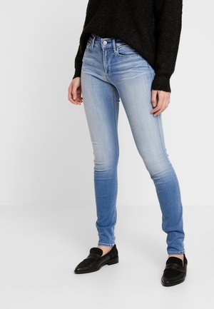 Persoon die lichtblauwe vervaagde skinny jeans, een zwarte trui en zwarte loafers draagt, staand op een effen witte achtergrond.