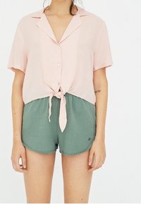 Short-sleeve light pink blouse with a tied front and buttons, paired with green elastic shorts featuring a small logo. Smooth texture.