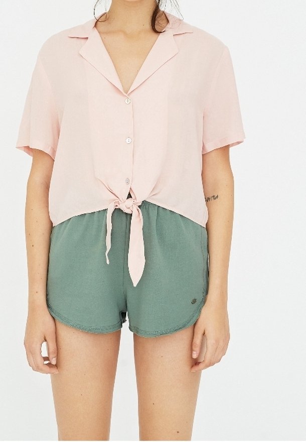 Short-sleeve light pink blouse with a tied front and buttons, paired with green elastic shorts featuring a small logo. Smooth texture.