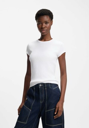 Young woman with short hair wears a fitted white t-shirt and dark blue wide-leg jeans against a plain white background.