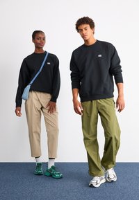 Kaks mudelit seisavad kõrvuti. Vasakul on seljas must kapuutsiga sviiter, beežid püksid ja rohelised tossud. Paremal on seljas must kapuutsiga sviiter ja oliivivärvi lahtised püksid.