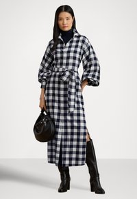 Pledēta krekla kleita tumši zilā un baltā krāsā ar apkakli, pogu aizdari priekšpusē, jostu pie vidukļa un balona formas piedurknēm. Melna rokas soma un ceļgalu garuma zābaki.