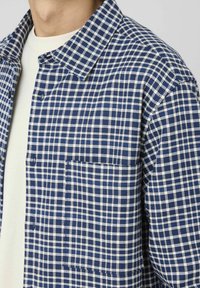 Chemise à carreaux bleus et blancs à boutons, fabriquée dans un tissu à tissage texturé, avec un col classique et une poche sur la poitrine.