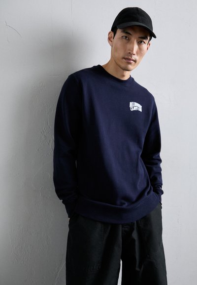 Mann iført en svart caps, marineblå sweatshirt med hvit "Billionaire Boys Club" tekst, og svarte bukser, stående mot en lys teksturert vegg.