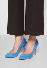 Blå mocka högklackade skor med spetsig tå och smal stiletthäl, tillsammans med svarta vida byxor.