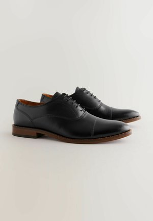 Zapatos de vestir negros de cuero con una textura suave, punta redondeada, detalles de costura sutiles y una suela de goma marrón.
