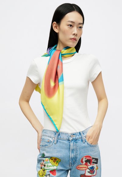 Femeie cu păr lung și închis la culoare, îmbrăcată cu o cămașă albă cu mâneci scurte, un fular colorat legat în jurul gâtului și blugi de un albastru deschis, cu mai multe broderii.