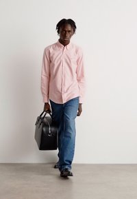 Camicia rosa a bottoni, jeans blu e borsa in pelle nera. Il modello ha un'espressione neutra e cammina su una superficie chiara contro uno sfondo semplice.