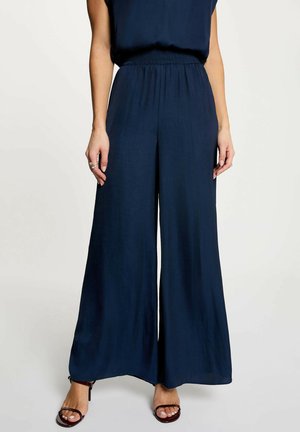 Femme portant un pantalon large bleu marine avec une ceinture élastique et des talons noirs à brides, debout devant un fond clair uni.