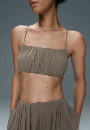 Femme portant un crop top taupe froncé avec fines bretelles et une jupe plissée assortie, les mains dans les poches, sur fond gris.