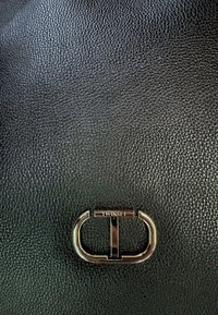 Pelle nera texturizzata con una centrale hardware ovale metallica che presenta l'incisione "TWINSET". Il design è minimalista e moderno.
