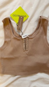 Brązowy prążkowany crop top z dekoltem w serek i zapięciem na napy z przodu. Metki wskazują na materiał z recyklingowanej bawełny. Tło to jasny materiał.