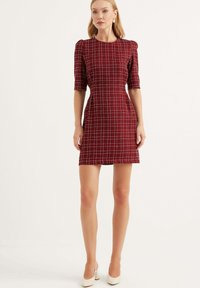 Röd rutigt klänning med en figursydd silhuett, korta puffärmar, knälång fåll och ett strukturerat tyg. Buren med vita högklackade skor.