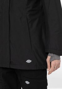 Svart vattentät jacka med rak skärning och slankt design, med en logopatch nära fållen och texturerade tygdetaljer.