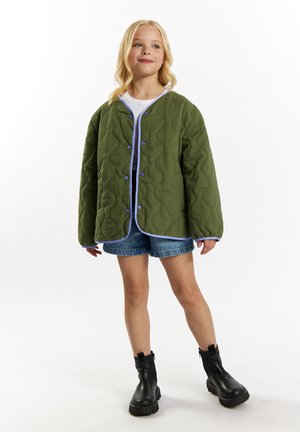Veste matelassée verte avec des accents bleus, à manches longues et fermetures à boutons, associée à un short en denim et des bottines noires.