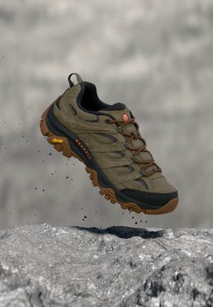 Chaussure de randonnée marron et noire avec une semelle robuste flottant au-dessus d'un sol rocailleux avec de petits cailloux qui tombent autour.
