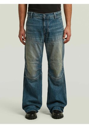 Homme portant un jean bleu ample avec des renforts aux genoux et des chaussures noires, debout devant un fond uni.