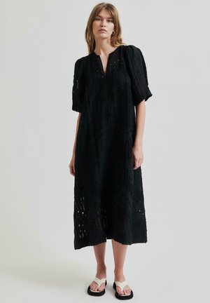 Black midi dress featuring a textured fabric with embroidered details, a V-neckline, and puffed sleeves. Paired with white sandals.