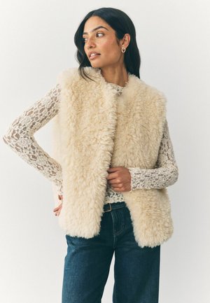 Faux-Fur-Weste in cremigem Beige mit flauschiger Textur. Getragen über einem langärmeligen Spitzenoberteil, mit dunkelblauen Jeans und sichtbarem Gürtel.