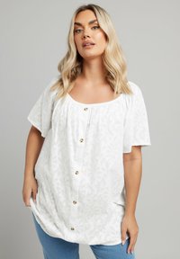 White floral-patterned blouse with short sleeves, square neckline, and decorative gold buttons down the front. Soft, textured fabric.