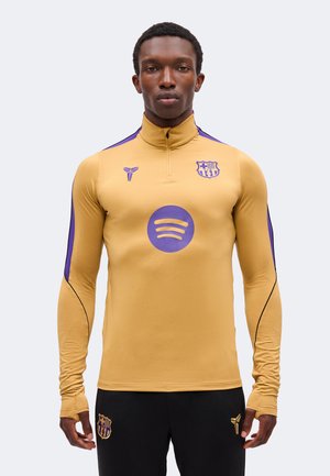 Mann trägt ein goldfarbenes und violettes Sportoberteil mit Reißverschluss sowie schwarze Hose, mit sichtbaren FC Barcelona- und Spotify-Logos auf der Kleidung.