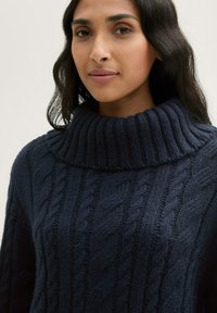 Navyblauer Kabelstrickpullover mit hohem geripptem Kragen. Verfügt über strukturierte Muster und lange Ärmel, hergestellt aus einem weichen, dicken Material.