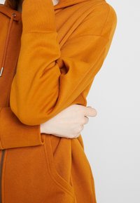 Sudadera naranja con cremallera, que cuenta con una capucha, bolsillos frontales y puños de canalé. La tela parece suave y texturizada.