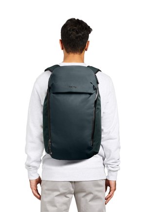 Homme portant un sac à dos Bellroy vert foncé avec des fermetures éclair verticales, vêtu d'un sweat-shirt gris clair et d'un pantalon beige, vu de dos sur fond blanc.