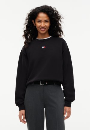 Kvinde med krøllet hår smiler iført en sort Tommy Jeans sweatshirt, grå bukser og et sort bælte mod en ensfarvet hvid baggrund.
