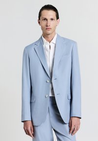 Junger Mann mit nach hinten gekämmtem Haar, der ein hellblaues Sakkos und eine Hose über einem weißen Hemd trägt, vor einem einfarbigen Hintergrund.
